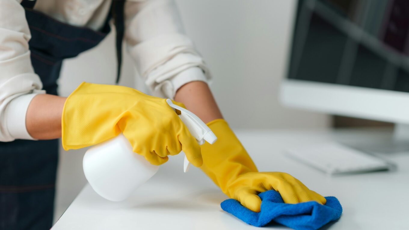 Close-up of hands in yellow gloves cleaning a white desk with a spray bottle and blue cloth