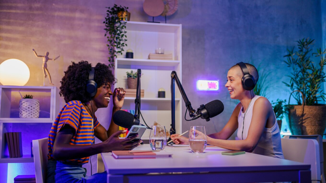 Two cheerful podcasters are sitting in a little home podcast studio.